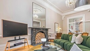 Ett modernt vardagsrum med en platt-TV, en öppen spis med vedeld, en grön soffa och en kristallkrona.