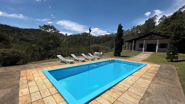 A rectangular swimming pool with a tiled deck, surrounded by white lounge chairs and a small building with a covered patio. - Rancho dos Capelos (Nazaré Paulista)