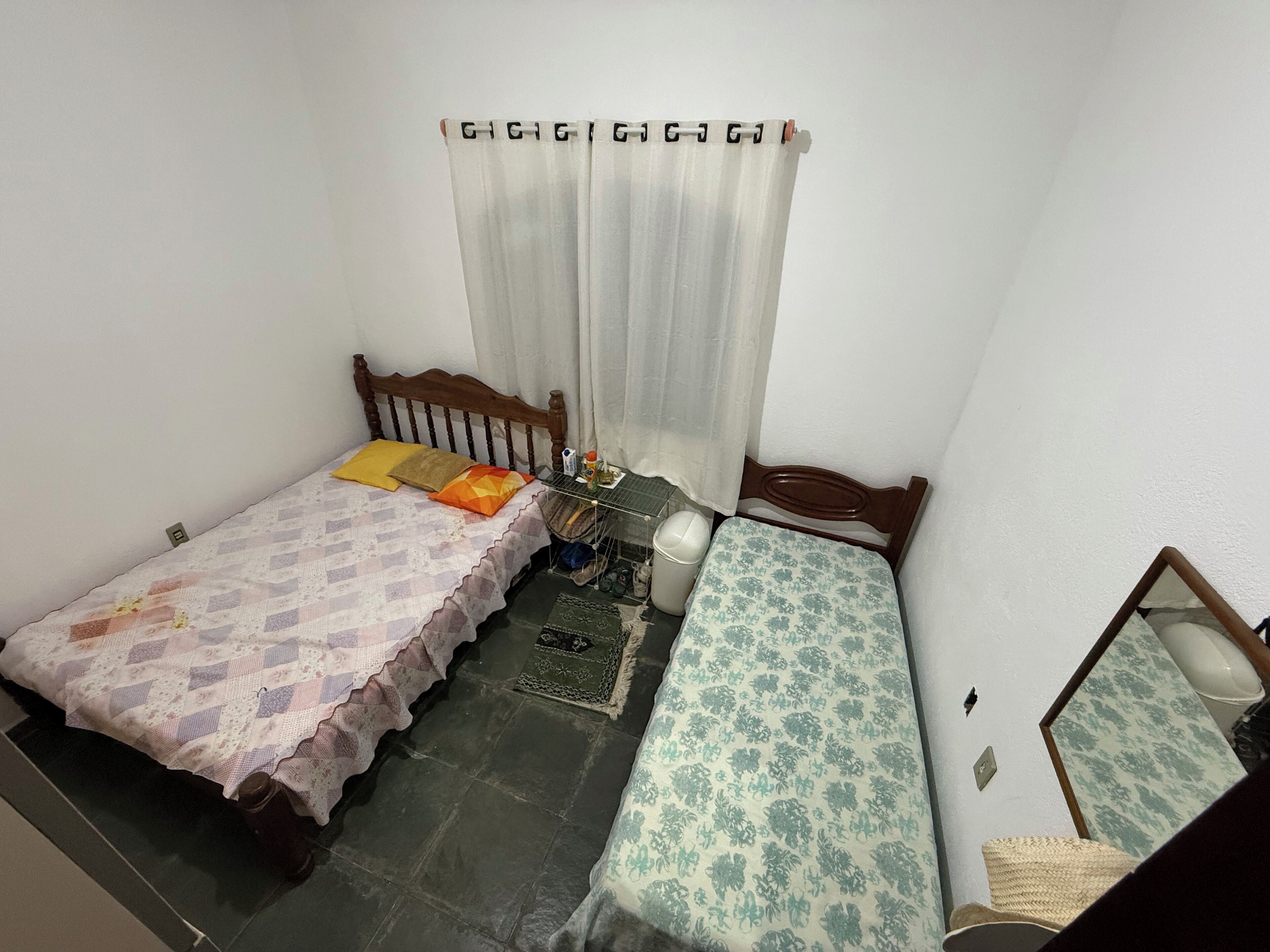 Quarto pequeno e compacto com duas camas de solteiro, cabeceira de madeira, uma mesinha e um espelho.