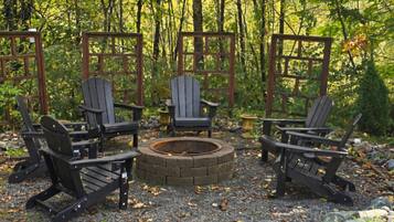 A circular fire pit surrounded by four Adirondack chairs, set on a gravel surface with a wooden fence and trees in the background.