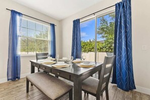 A dining area with a table set for four, large windows with blue curtains, and a view of greenery outside. - Pool * Game Room * Mins to Disney (Kissimmee)
