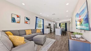 A modern living room with a grey sectional sofa, a kitchen in the background, and a large TV on the wall. - Pool * Game Room * Mins to Disney (Kissimmee)