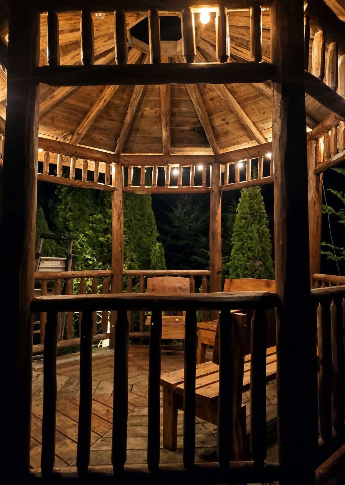 A wooden gazebo with a roof, surrounded by a wooden deck and chairs.