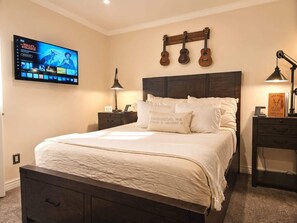 Un dormitorio con cama, televisor montado en la pared, mesita de noche, lámpara y un soporte para guitarras en la pared.