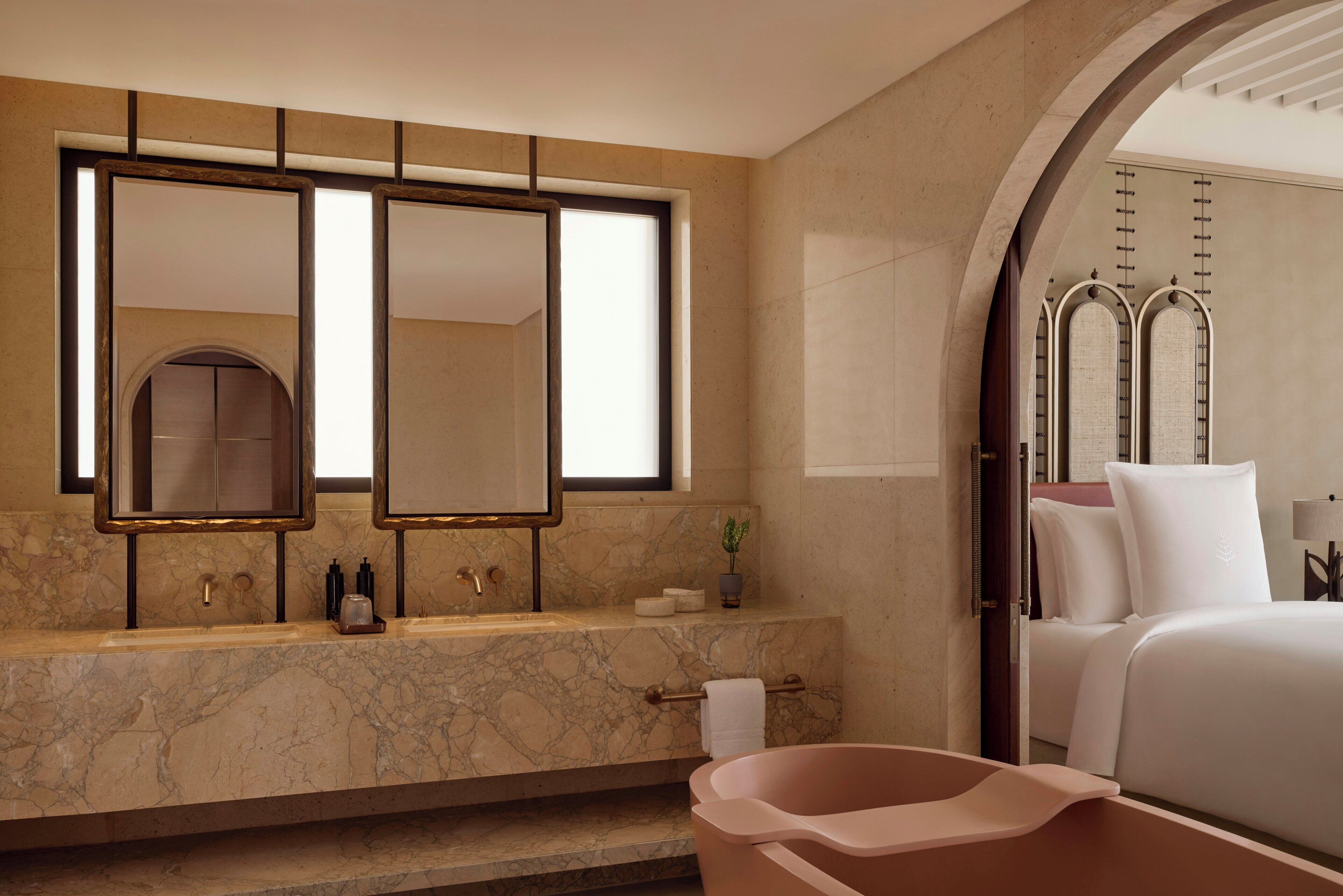 A modern bathroom with a marble sink, dual mirrors, a bathtub, and a bed with white linens.