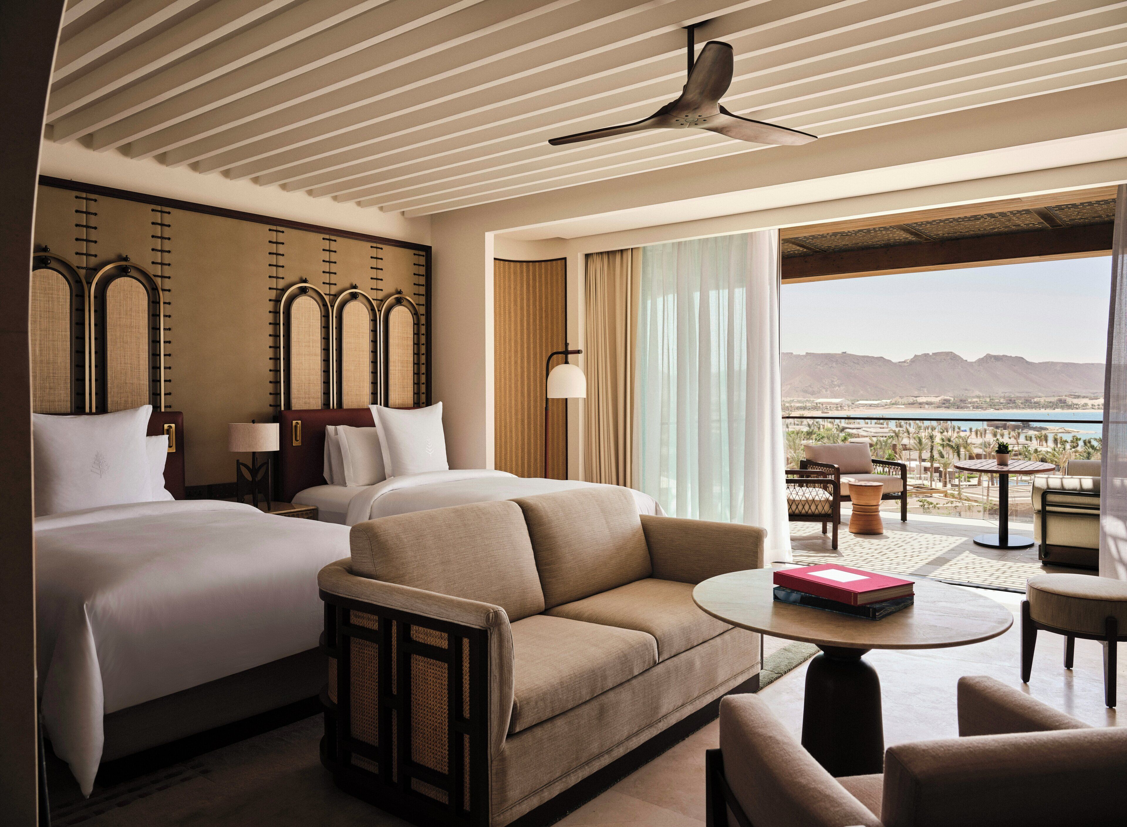 A hotel room with a bed, a sofa, a table, and a view of the mountains and sea.
