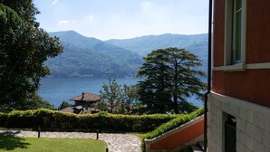 Vista di un lago con montagne sullo sfondo, un edificio con facciata rossa e un sentiero in pietra che conduce a una casa con tetto in tegole.