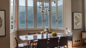 Zona pranzo con un grande tavolo, sedie, lampade a sospensione e vista su un lago attraverso ampie finestre.