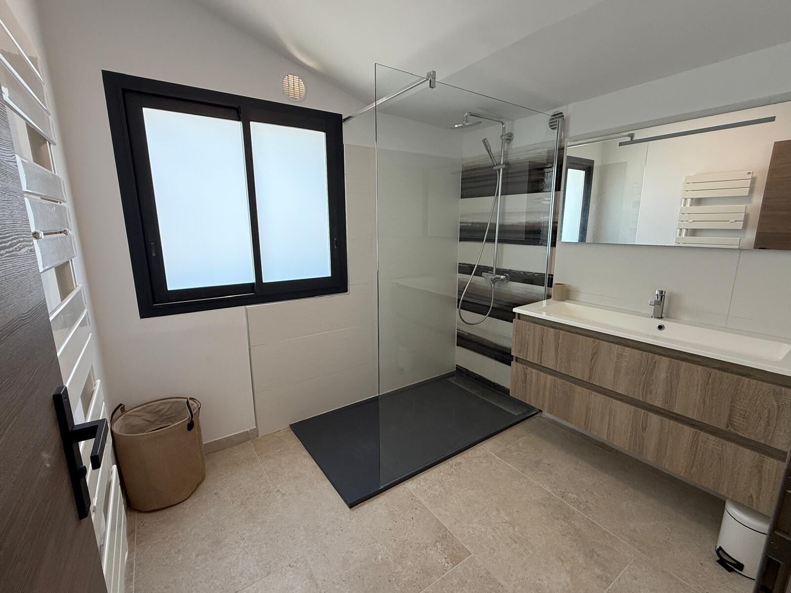 A modern bathroom with a glass shower enclosure, a large window, and a vanity with a mirror.