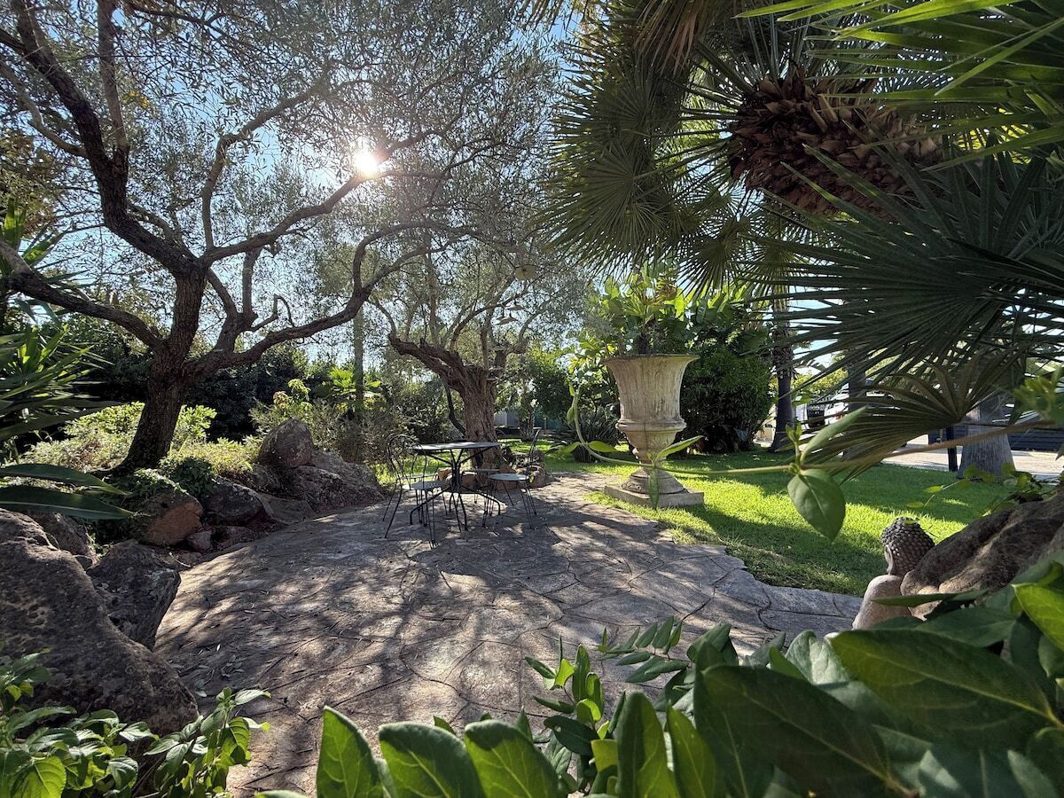 Un espace patio avec une table et des chaises en pierre, entouré d’une végétation luxuriante et agrémenté d’un grand bac à fleurs.