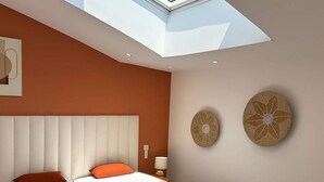 Un dormitorio con tragaluces, una cama con ropa de cama blanca y naranja, dos elementos decorativos en la pared y un cabecero de color claro.