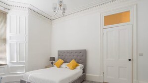 A bedroom with a grey upholstered bed, white walls, and a white door. - Main Door Private Entrance Apartment (Edinburgh)