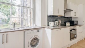 A modern kitchen with white cabinets, a built-in washing machine, a sink, a toaster, a microwave, and a view of the outside through large windows. - Main Door Private Entrance Apartment (Edinburgh)