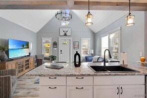 A spacious kitchen with a granite countertop, white cabinetry, and a large island with a sink. There is a chandelier, a television, and a dining area visible. - *Perfect Vacation* Cozy Lancaster Village Bungalow (Lancaster)