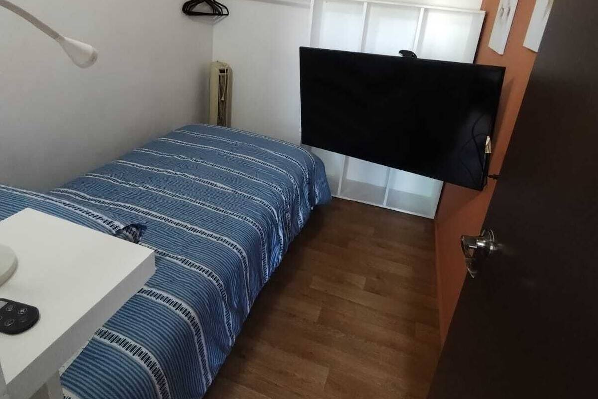 A small, compact room with a single bed, a desk, a TV mounted on the wall, and a door.