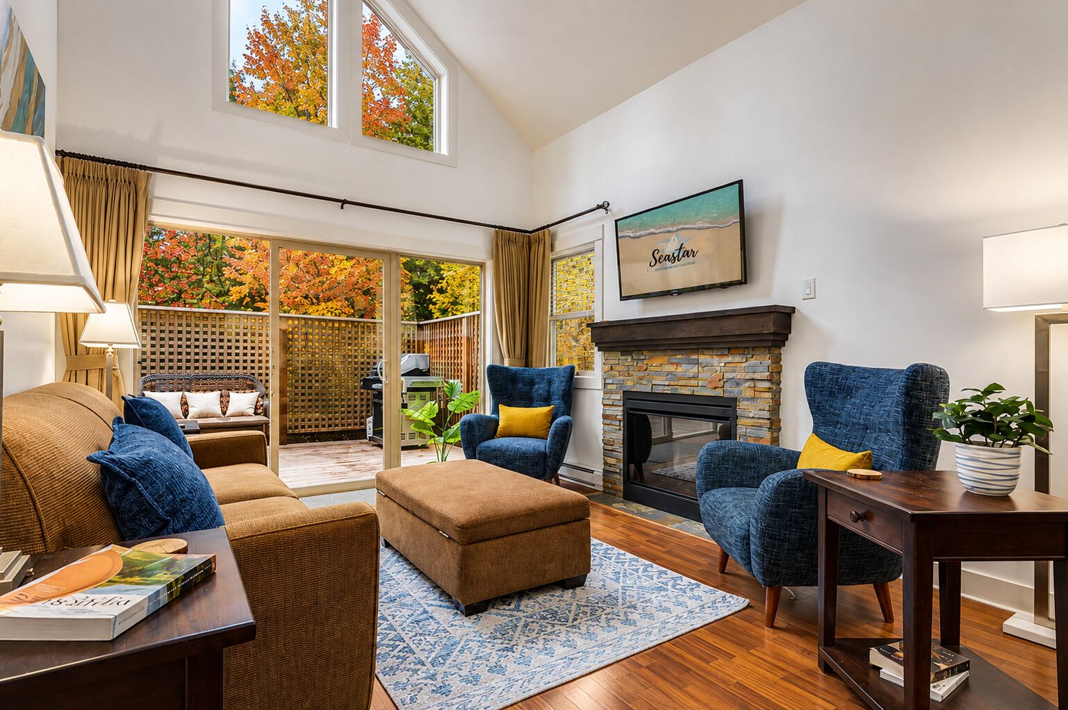 A living room with a fireplace, blue armchairs, a brown sofa, and a large window with autumn foliage.