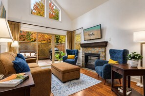 A living room with a fireplace, blue armchairs, a brown sofa, and a large window with autumn foliage.