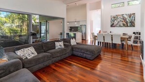 Un salon moderne avec un grand canapé d’angle, un coin repas avec une table et des chaises blanches, et une cuisine en arrière-plan.