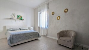 Un dormitorio con cama, una silla, un ventanal con cortinas y adornos en la pared.
