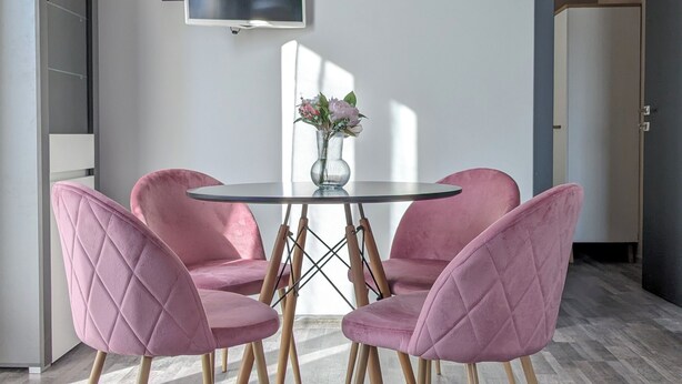 Un comedor con una mesa de vidrio, cuatro sillas tapizadas en rosa, un jarrón con flores y un televisor de pantalla plana montado en la pared.
