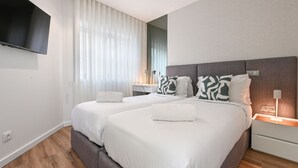 A modern hotel room with two beds, a flat-screen TV mounted on the wall, a large window with sheer curtains, and a bedside table with a lamp. - ON Apartments - Matosinhos Premium  (Matosinhos)