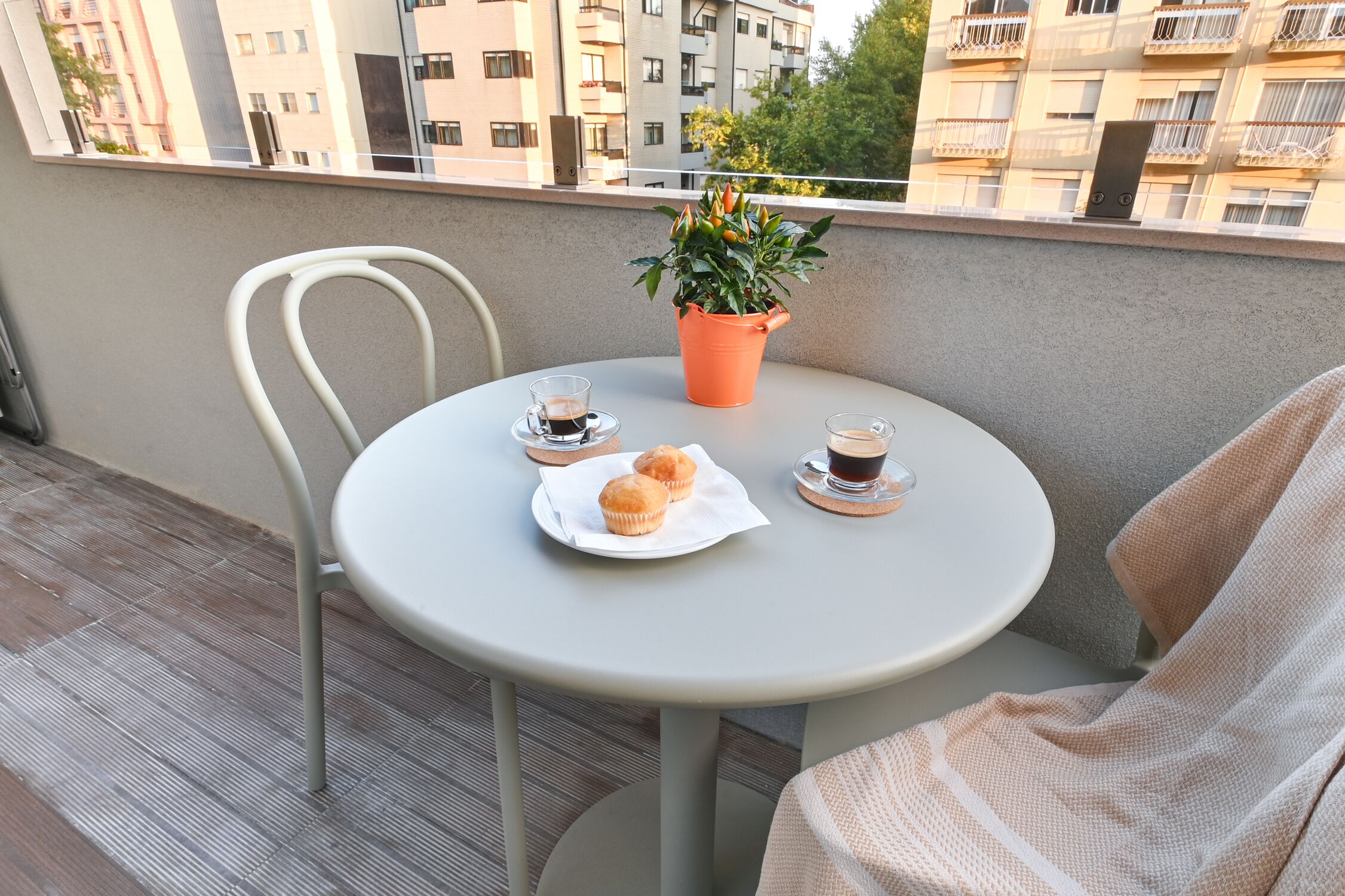 Ein runder Tisch mit einem weißen Stuhl, zwei Kaffeetassen, einem Tablett mit Muffins und einem Blumentopf auf einem Balkon mit Blick auf Wohnhäuser.