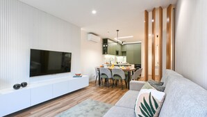 A modern living room with a flat-screen TV mounted on a white wall, a grey sofa with decorative pillows, a dining area with a wooden table and chairs, and a kitchen visible through an open door. - ON Apartments - Matosinhos Premium  (Matosinhos)