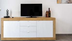 Een moderne woonkamer met een flatscreen televisie op een houten console, een schilderij aan de muur en decoratieve flessen.