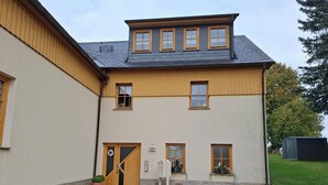 A two-story building with a sloped roof, a wooden door, and a small porch area. - Vacation apartment Am Ranis (Oederan)