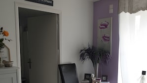 Een kamer met een zwarte stoel, een tafeltje met een vaas en fotolijstjes, een bord boven de deur en een roze vloerkleed.