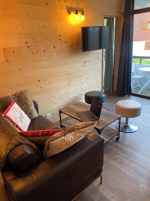 Ein gemütliches Zimmer mit einer Ledersofa, einem Laptop auf einem Holztisch, einem an der Wand befestigten Fernseher und Blick ins Freie durch eine Glastür.