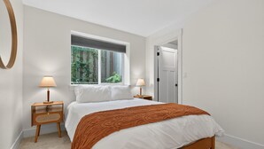 A bedroom with a large bed, two bedside tables with lamps, a window with blinds, and a door. - St. Johns Nest, Near Cathedral Park (Portland)