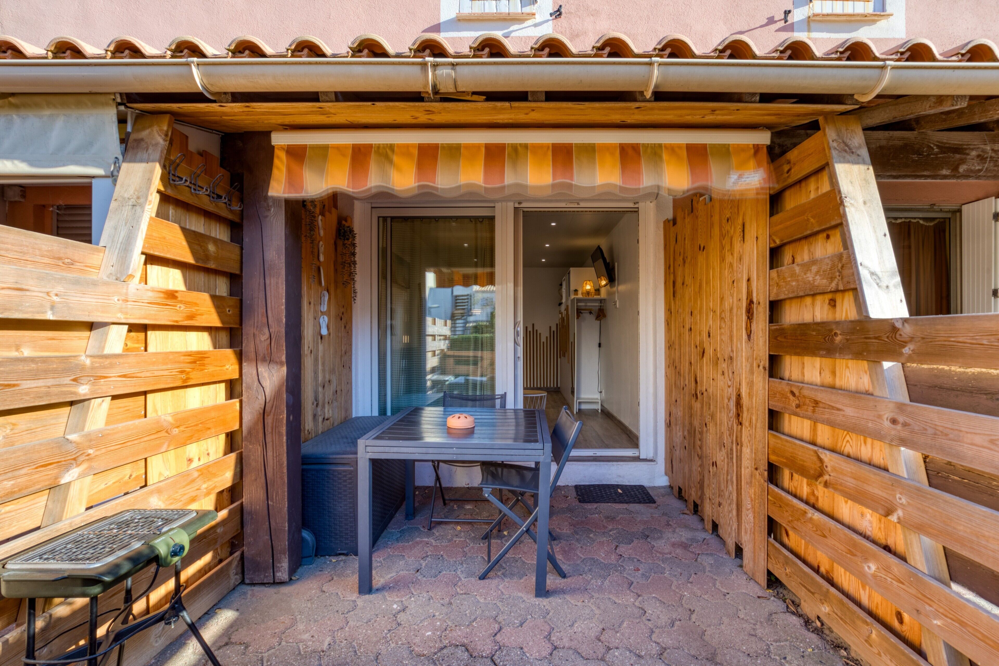 Une terrasse extérieure en bois, avec une table et des chaises, une petite table avec un clavier et un auvent à rayures.