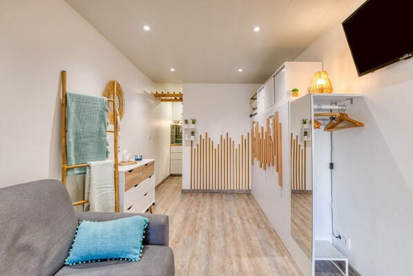 A modern room with a grey sofa, a wooden floor, a kitchen area, and a TV mounted on the wall. - Cozy Studio with Private Terrace – Ideal for a Seaside Getaway (Cap d'Agde)