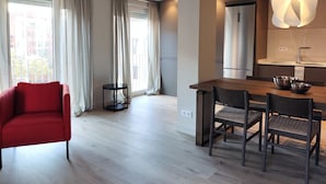 Ein modernes Wohnzimmer mit einem roten Sessel, einem Esstisch mit Stühlen, einer Küchenzeile mit Kühlschrank und Spüle sowie großen Fenstern mit Vorhängen.