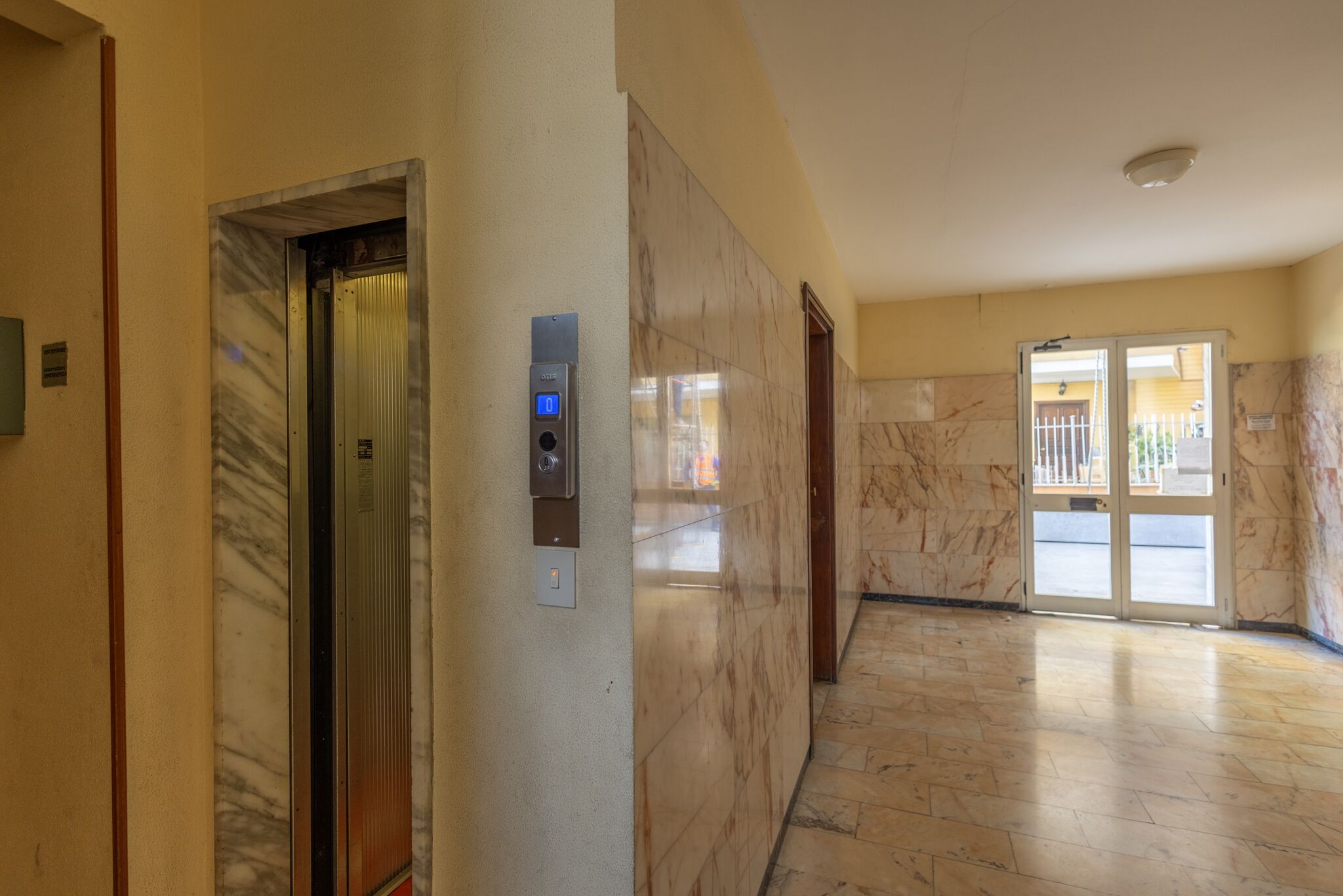 Un pasillo con paredes de mármol, un ascensor y una puerta que da a un espacio al aire libre.