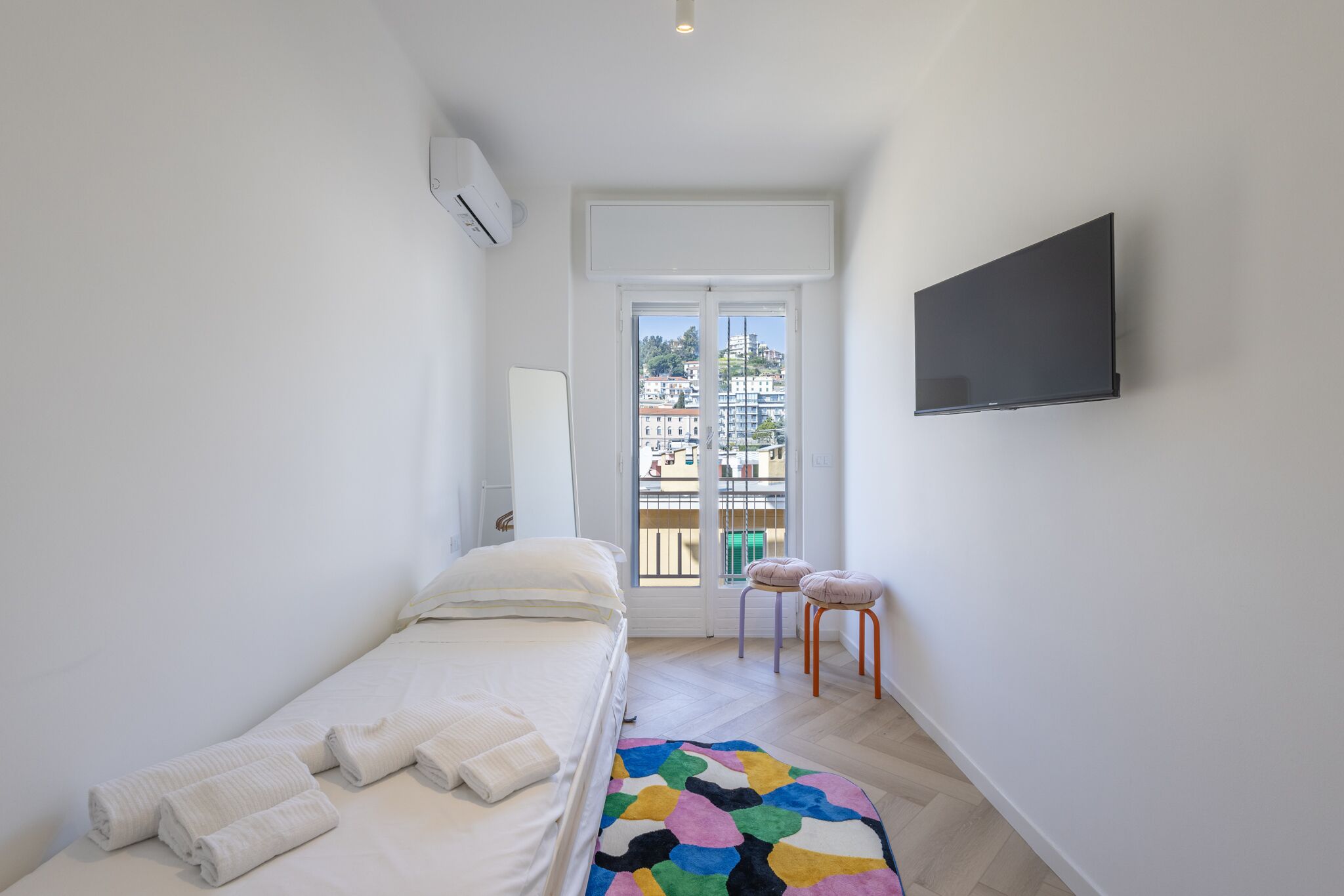 Una habitación pequeña y luminosa con una cama, un tapete colorido, un televisor y vistas a edificios por una puerta.
