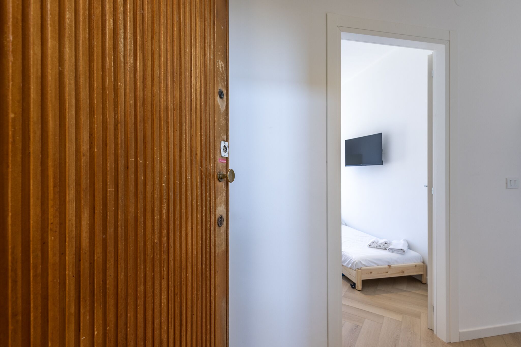 A room with a wooden door, a bed with a mattress and pillows, a flat-screen TV mounted on the wall, and a light-colored floor.