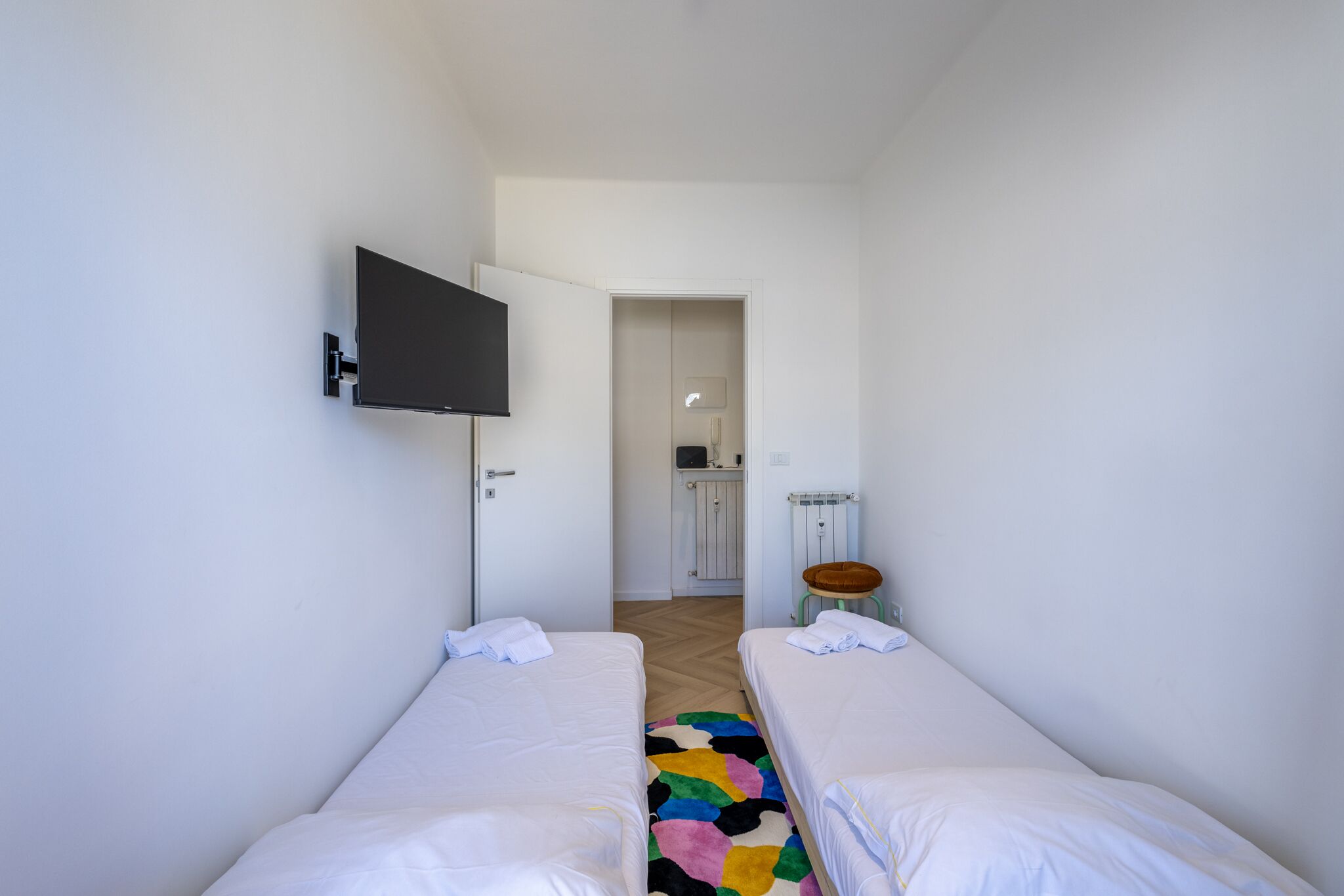 Une chambre d’hôtel avec deux lits simples, un téléviseur fixé au mur, un petit tabouret et une porte donnant sur une salle de bain.