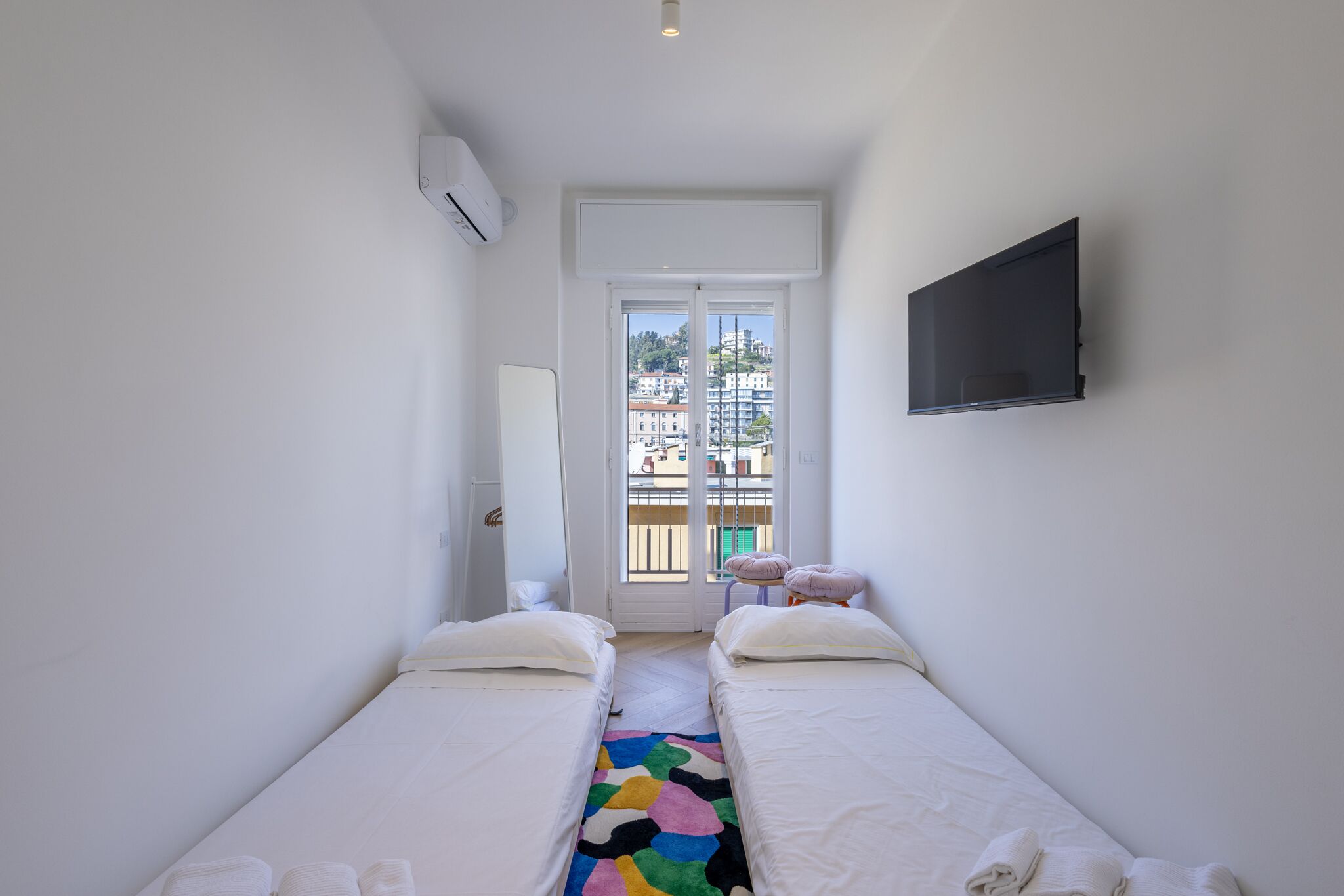 Une petite chambre lumineuse, avec deux lits simples, un téléviseur fixé au mur et une fenêtre donnant sur des immeubles.