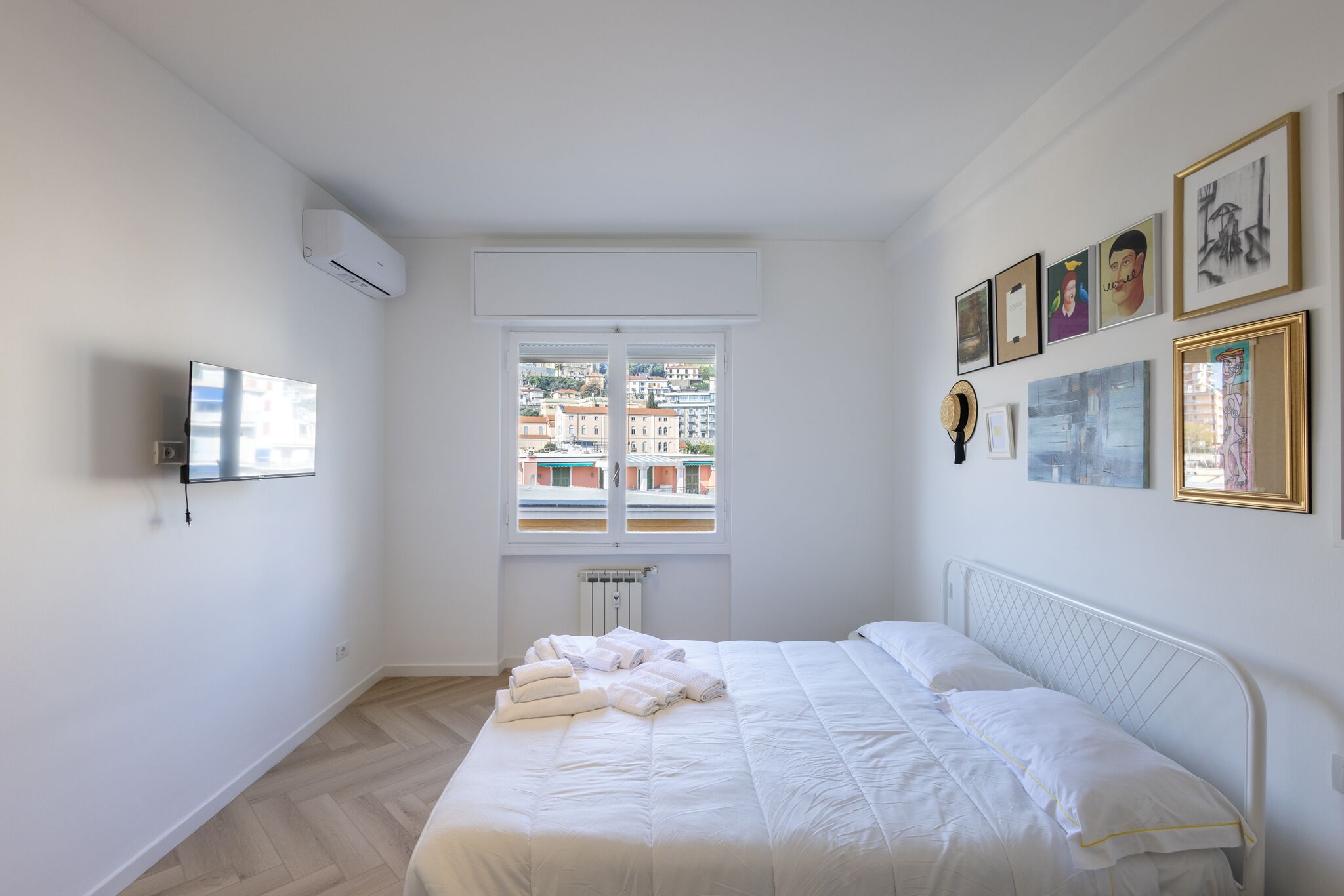 Ein Schlafzimmer mit Bett, Fenster mit Blick auf Gebäude, einem montierten Fernseher und gerahmten Kunstwerken an der Wand.