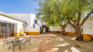 Une cour avec un sol carrelé, une table ronde entourée de chaises et un grand arbre qui offre de l’ombre.