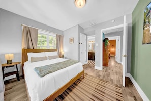 A bedroom with a wooden bed, a nightstand, a window with curtains, and a hallway leading to a bathroom.
