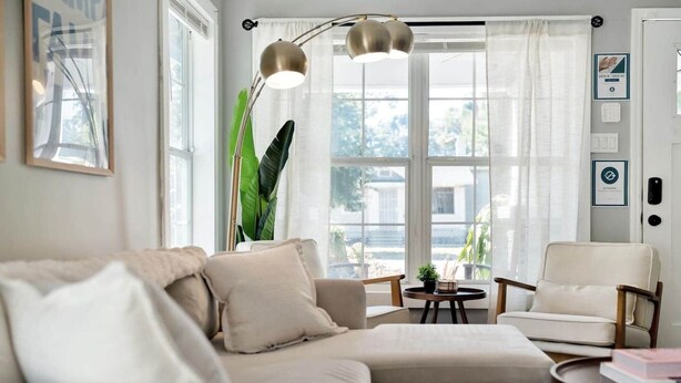 Ett vardagsrum med en soffa, fåtöljer, ett soffbord, en golvlampa och ett stort fönster med gardiner.