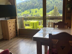 A room with a wooden dresser, a table with a plastic cover, a yellow chair, and a balcony with a view of a green landscape.