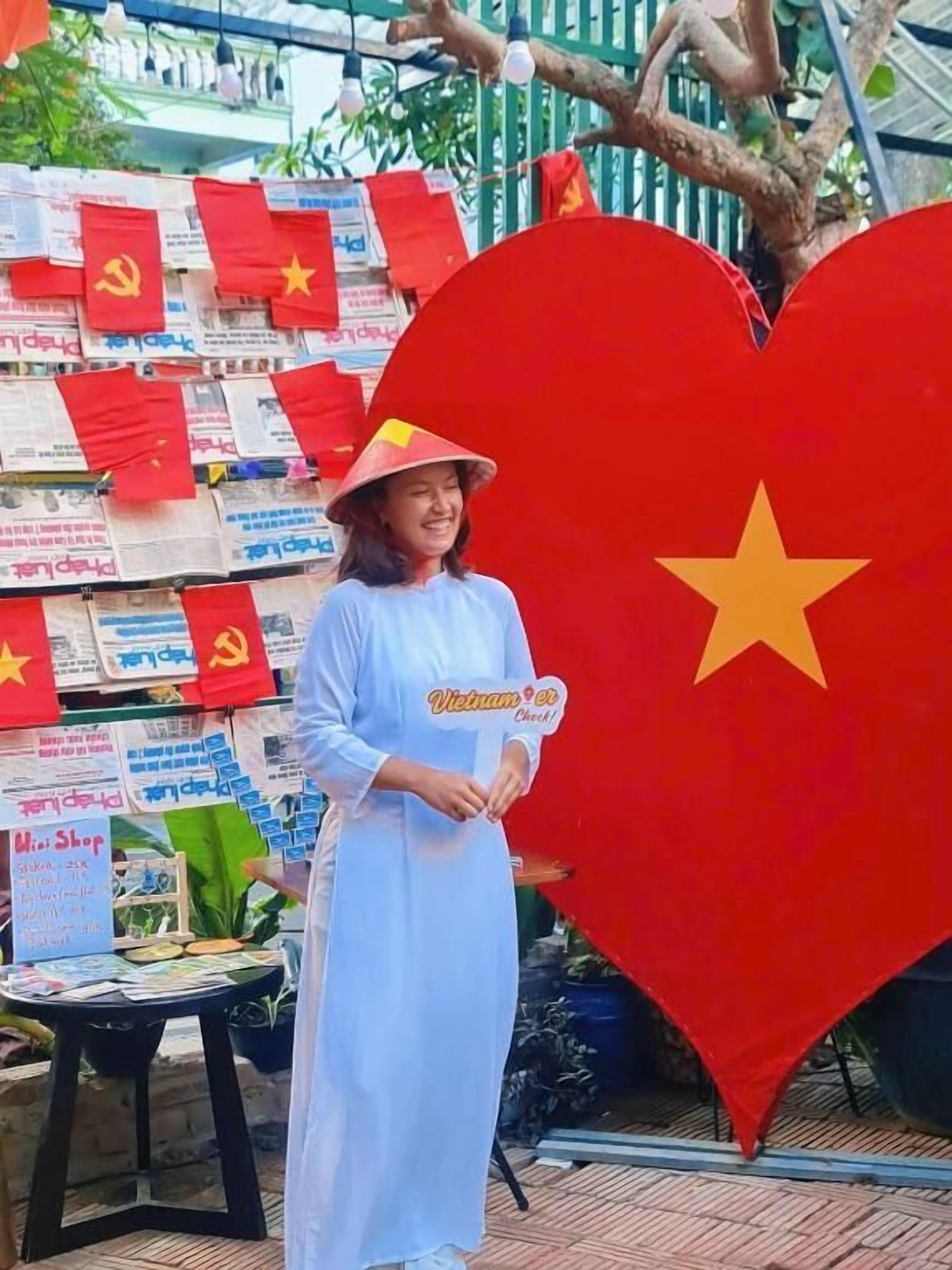 Une personne se tient devant un grand drapeau en forme de cœur orné d’une étoile, entourée de plus petits drapeaux et d’une enseigne sur laquelle on peut lire « Mini Shop ».