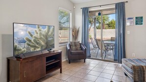 Ein Wohnzimmer mit Flachbildfernseher, einer dunkelholz-Unterhaltungseinheit, einem grauen Sessel, einem Essbereich mit Tisch und Stühlen sowie einer Glastür, die zu einer Außenpatio führt.