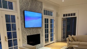 En rummelig stue med et fladskærms-tv monteret på en marmorvæg, en pejs og et stort vindue med glasdøre.
