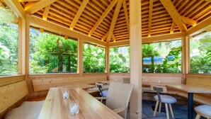A wooden pavilion with benches and tables, large windows, and a view of greenery.