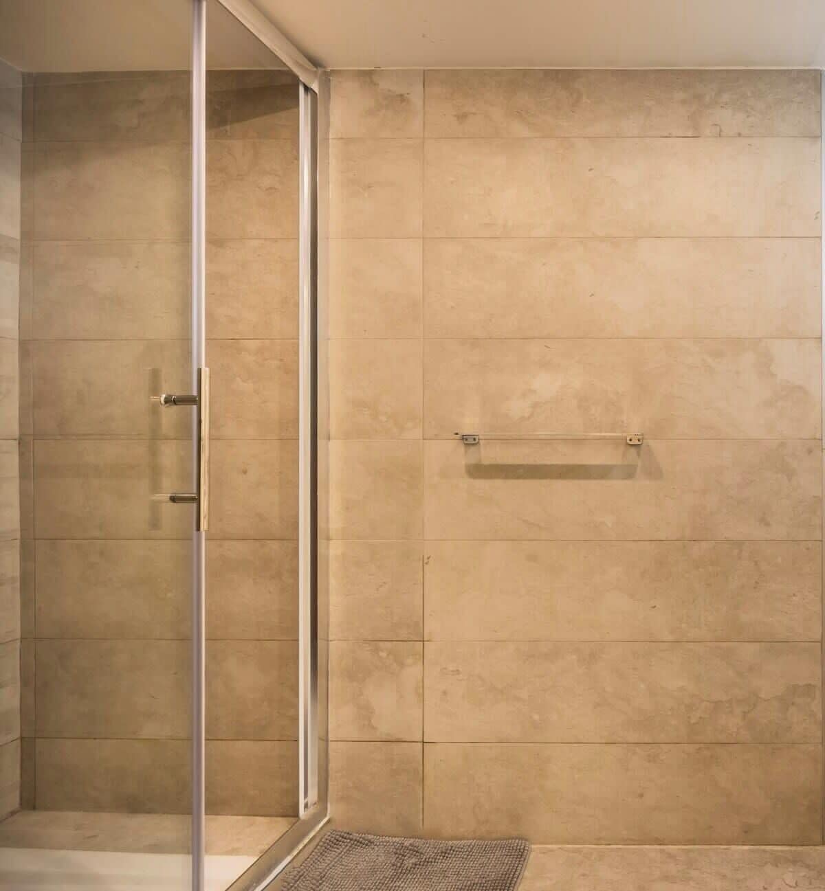 Zona de ducha con puerta de vidrio, azulejos color beige y un perchero para toallas.